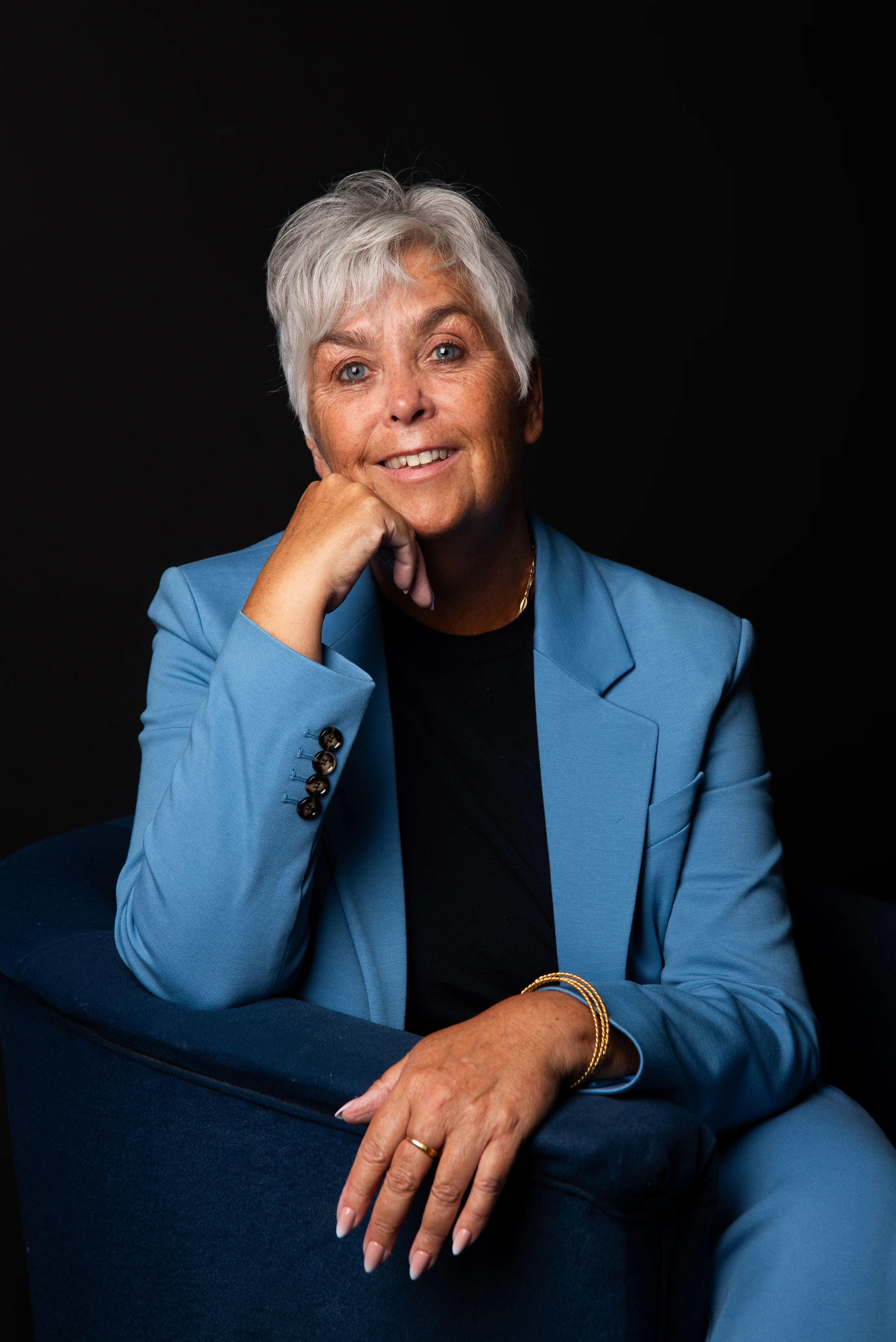 Arlette Vonk in a light blue suit, seated in an armchair against a dark backdrop, looking into the camera